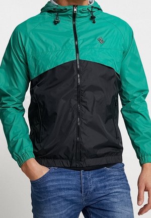 Mężczyzna w bluzie z kapturem typu windbreaker, z zieloną górną i czarną dolną częścią, zapinanej na suwak, z rękami w kieszeniach niebieskich dżinsów.