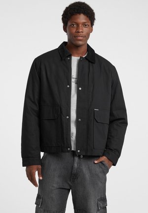 Giubbotto nero con colletto, due tasche frontali e chiusure a pressione; indossato sopra una camicia grigia; texture liscia e design semplice.