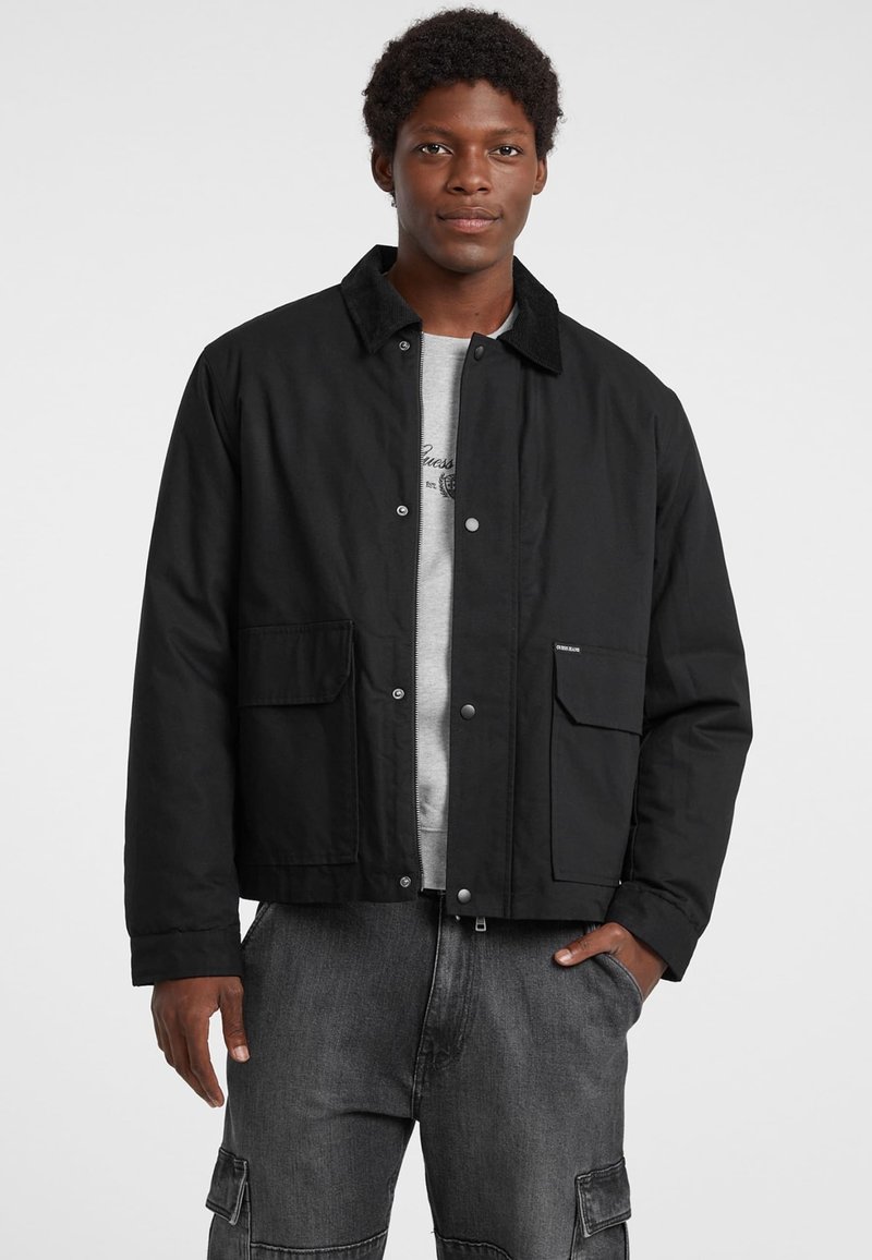 Giubbotto nero con colletto, due tasche frontali e chiusure a pressione; indossato sopra una camicia grigia; texture liscia e design semplice.
