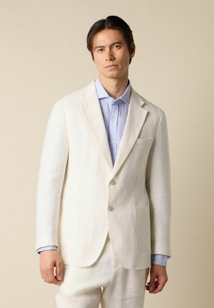 Junger Mann trägt ein cremefarbenes Sakko und eine Hose mit einem blau gestreiften Hemd vor einem beigen Hintergrund.