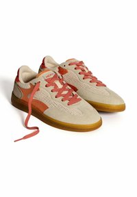 Zapatillas de ante beige con acentos de malla, detalles texturizados en rojo y branding naranja. Presentan suelas de goma gruesas y cordones a juego.