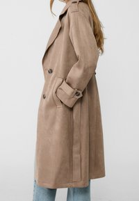 Beige suède trenchcoat met dubbele knopen, schouderepauletten en zijzakken, gecombineerd met lichte denim jeans.