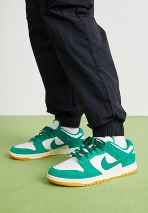 Baskets Nike blanches et vertes portées avec un pantalon noir et des chaussettes blanches sur un sol vert avec un arrière-plan gris clair.