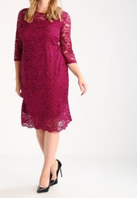 Burgundy spetsklänning med trekvartsärmar, blommönster, scallopad fåll, i kombination med svarta högklackade skor. Slät textur.