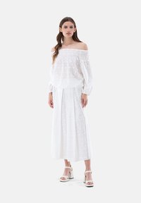 Weiße schulterfreie Bluse mit Raffung am Ausschnitt, kombiniert mit einem langen weißen Rock. Das Outfit hat ein strukturiertes Blumenmuster.
