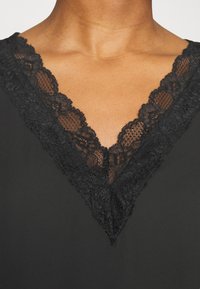Vestuário preto com um design em V, adornado com um intrincado acabamento de renda preta ao longo da gola, criando um contraste texturizado com o tecido.