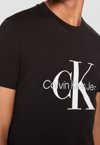 Primer plano de una persona que lleva una camiseta negra con un gran logo blanco "CK" y un logo gris de "Calvin Klein Jeans" en el pecho.
