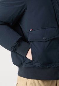 Marineblauwe gewatteerde jas met een voorflapzak met een decoratieve driekleurige accent, elastische manchetten en een getailleerde tailleband.