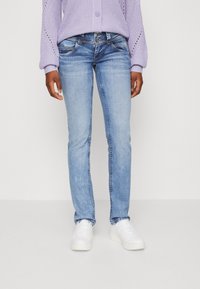 Personne portant un jean en denim bleu clair, des baskets blanches et un pull en tricot lavande, se tenant devant un fond uni.