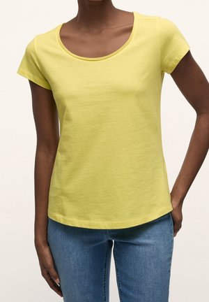 Femme portant un t-shirt jaune à manches courtes et un jean bleu, debout avec une main dans la poche sur un fond uni.