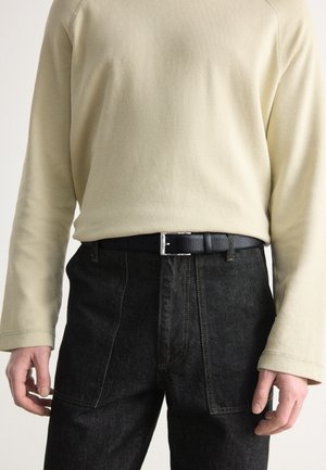 Cintura in pelle nera con fibbia argentata, indossata sopra pantaloni in denim scuri e una camicia a maniche lunghe in beige chiaro con texture e polsini a coste.
