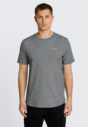 Mężczyzna noszący prostą szarą koszulkę z krótkim rękawem z małym napisem „Denim Design Studio” na piersi, w połączeniu z czarnymi spodniami, neutralne tło.