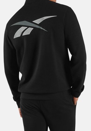 Person, der einen schwarzen Pullover und schwarze Hose mit einem großen silbernen und grauen abstrakten Logo auf dem oberen Rücken trägt, steht mit den Händen in den Taschen.