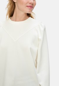Weißer Pullover mit einem runden Ausschnitt und einem V-förmigen Schulterdetails. Mit Puffärmeln und einer glatten, weichen Textur.