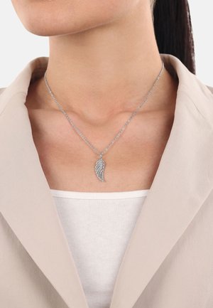 Silberne Halskette mit einem Flügel-Anhänger, der filigrane Details aufweist. Über einem weißen Oberteil und unter einem beigen Blazer getragen.