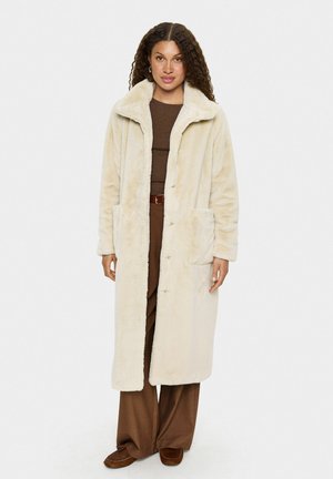Langer, cremefarbener Fleece-Mantel mit weicher Textur, umgeschlagenem Kragen und zwei Fronttaschen. Getragen über braunen Hosen und einem Oberteil.