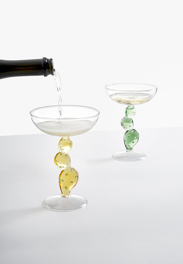 Szampan nalewany do kieliszka coupe z żółtą teksturowaną nóżką, obok podobnego kieliszka z zieloną teksturowaną nóżką.