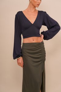 Navy kék, hosszú ujjú crop top, mély V-kivágással és redőzött ujjakkal, párosítva egy magas derekú, olíva zöld szoknyával, amelyen redőzött rész és oldalhasíték található.