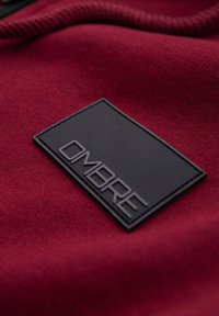 Étiquette de marque rectangulaire noire avec texte en relief « OMERE » sur une surface en tissu rouge foncé texturée.