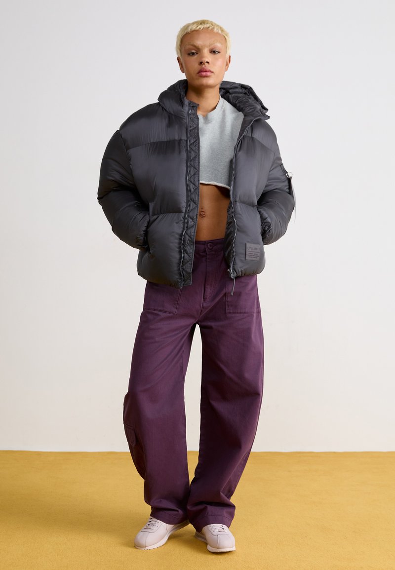 Chaqueta tipo puff en gris oscuro, de diseño corto, con capucha y costuras horizontales; combinada con pantalones morados de pierna ancha y zapatos claros.