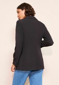 Juodas švarkas, pagamintas iš lygaus audinio, su klasikiniais apykaklės kraštais ir pritaikytu siluetu, derinamas su šviesiai mėlynomis džinsinėmis kelnėmis.