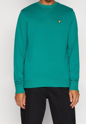 Hombre que lleva una sudadera de manga larga color verde azulado con un pequeño logo de un pájaro amarillo en el pecho izquierdo y pantalones negros.