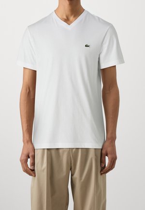 Mand iført en enkel hvid V-hals T-shirt med et lille grønt krokodillelogo på venstre bryst, kombineret med beige bukser.