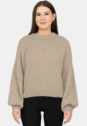Jonge vrouw met lang donker haar draagt een beige grofgebreide trui en zwarte broek, staand tegen een effen lichte achtergrond.
