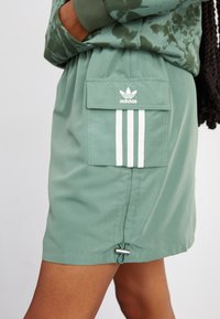 Zöld sportnadrág rugalmas derékkal, fehér csíkkal és Adidas logós zsebből áll. Sima anyag matt felülettel.