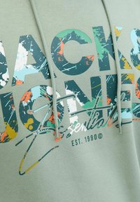 Olijfgroene hoodie met grote, multikleurige grafische tekst. Materiaal: zachte katoenmix. Treksnoer aan de capuchon. Getextureerd oppervlak met spetters.