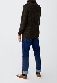 Pull texturé vert foncé avec un col montant, associé à un jean bleu avec les revers retroussés et des baskets marron. Look simple et décontracté.