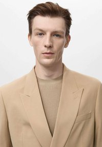 Blazer beige realizzato in tessuto liscio con rever affilati, indossato sopra un maglione beige a trama. Linee pulite, dettagli minimi e vestibilità moderna.