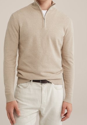 Beiger, strukturierter Strickpullover mit Viertel-Reißverschluss, getragen über einem weißen Hemd, kombiniert mit cremefarbenen Hosen und dunkelbraunem Gürtel.