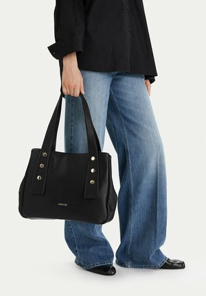 Persona che indossa jeans blu a gamba larga e scarpe nere, che tiene in mano una borsa tote nera in pelle con borchie metalliche e logo LASOCKI.