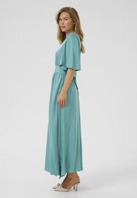 A long teal dress with short, wide sleeves, a cinched waist, and a flowing silhouette. Paired with beige heeled mules. Smooth texture.