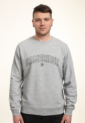 Jeune homme portant un sweat-shirt gris avec le texte "CAMBRIDGE" en grandes lettres, debout devant un fond clair uni.