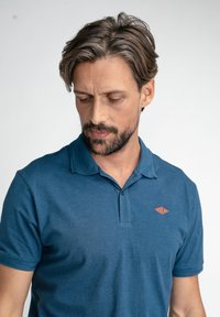 Camisa de polo a rayas azules con un cuello corto, que presenta un solo botón y un acento de logo naranja en el lado izquierdo del pecho. Textura suave.