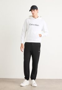 Hanorac alb cu glugă cu logo-ul "Calvin Klein", asortat cu pantaloni joggeri negri cu un logo mic și adidași albi. Se poartă o șapcă neagră.