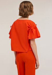 Blusa di cotone rossa con maniche corte a volant e dettaglio con cordoncino, caratterizzata da un taglio morbido. Abbinata a pantaloni rossi coordinati.