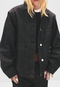 Svart denimjacka med avslappnad passform, med knappstängning, bröstficka och synlig sömdekor. Bärs över en svart topp.