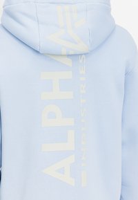 Világoskék kapucnis pulcsi, puha tapintással. A hátoldalán egy nagy, krémszínű grafikai szöveg látható, amely függőlegesen van nyomtatva: 'ALPHA INDUSTRIES'.