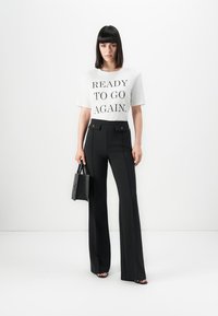 Woman stands wearing white t-shirt with "READY TO GO AGAIN" text, black high-waisted pants, black sandals, holding small black handbag.