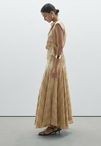 Langes, fließendes Kleid aus einem hellen, tanfarbigen Stoff mit einem subtilen Karomuster. Verfügt über einen drapierten Ausschnitt und minimalistische schwarze Sandalen.