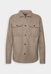 Veste-chemise en laine beige avec deux poches poitrine à rabat, manches longues et col classique sur un fond uni.