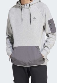 Šedá mikina s kapucí a černým spodním panelem, která má přední kapsu, nastavitelné šňůrky a logo Adidas na hrudi. Boční zipová kapsa na rukávu.
