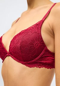 Κοντινό πλάνο μιας γυναίκας που φοράει ένα κόκκινο φλοράλ δαντελένιο σουτιέν με λεπτές τιράντες και ενίσχυση κάτω από το στήθος, σε απλό φόντο.