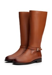 Brune skinn overkneboots med en glatt finish, med en dekorativ spenne stropp og en rund tå, satt på en lav trerand.