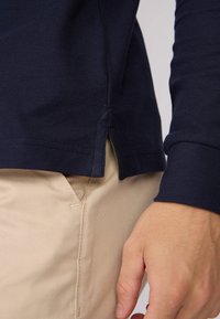 Chemise à manches longues bleu marine en tissu texturé, avec un ourlet fendu et des poignets. Associée à un pantalon beige clair.