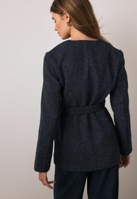 Granatowa teksturowana kurtka z paskiem w talii, długimi rękawami i prostym krojem. Charakteryzuje się minimalistycznym designem i gładkim wykończeniem.