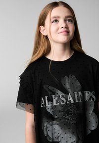 Melna T-kreklu ar punktiņu tekstūru, kurā redzams ziedu grafiskais attēls un vārds "ALLSAINTS" baltā krāsā. Viensrūpju akcents īsajās piedurknēs.